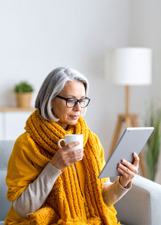 Femme âgée lisant une tablette, tasse à la main.